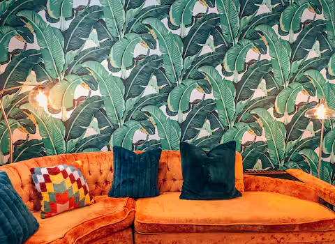 Bright orange mid-century sofa with colorful pillows against tropical banana leaf wallpaper at vacation rental in Panama City Beach.