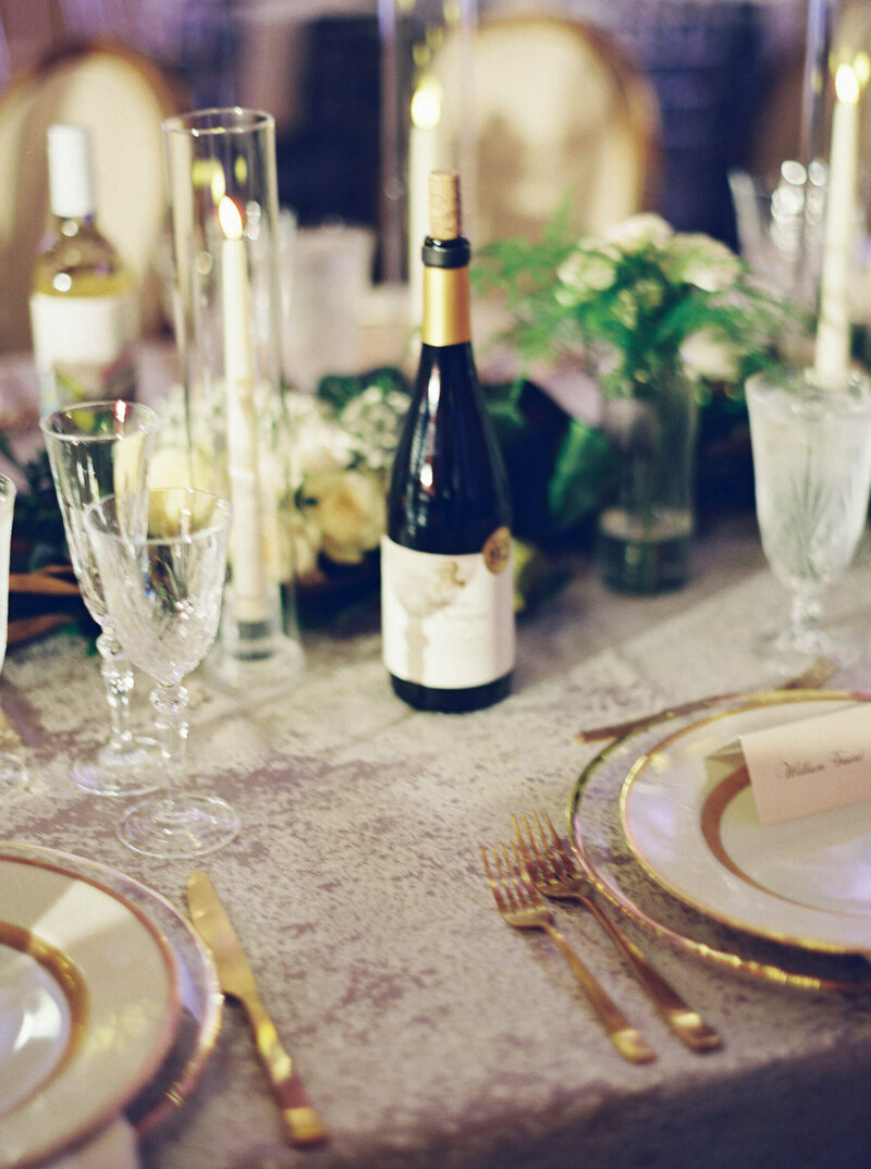 A picture of a bottle of wine on a table with linen covers and white florals the table is decorated for the wedding reception
