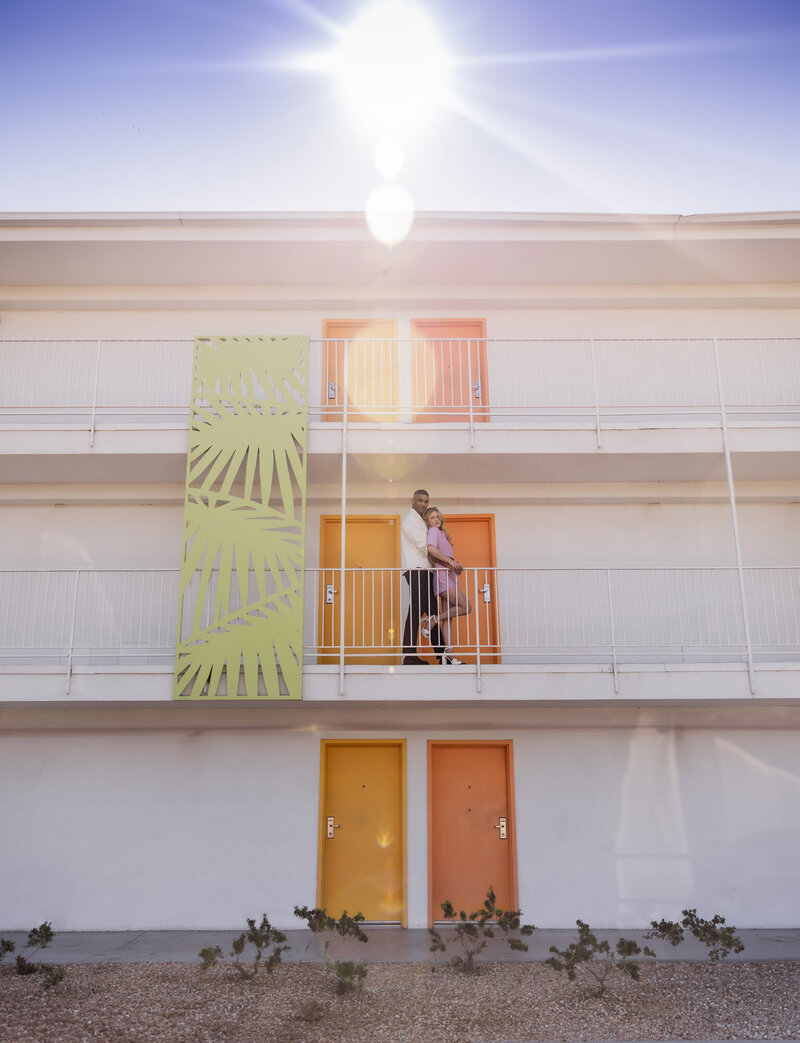 A stylish couple embraces on a balcony in Palm Springs, California, with bold pops of color from the retro-modern architecture. The sun flares overhead, creating a dreamy and cinematic vibe that captures the essence of desert romance and vibrant design.