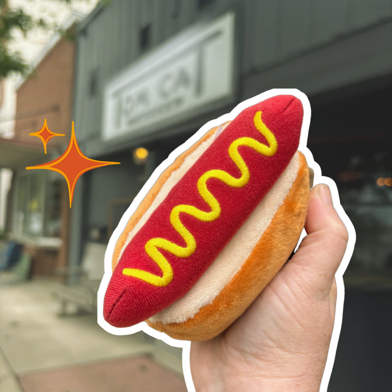 Hand holding a plush hot dog dog toy in front of a pet store, highlighted with sparkles.