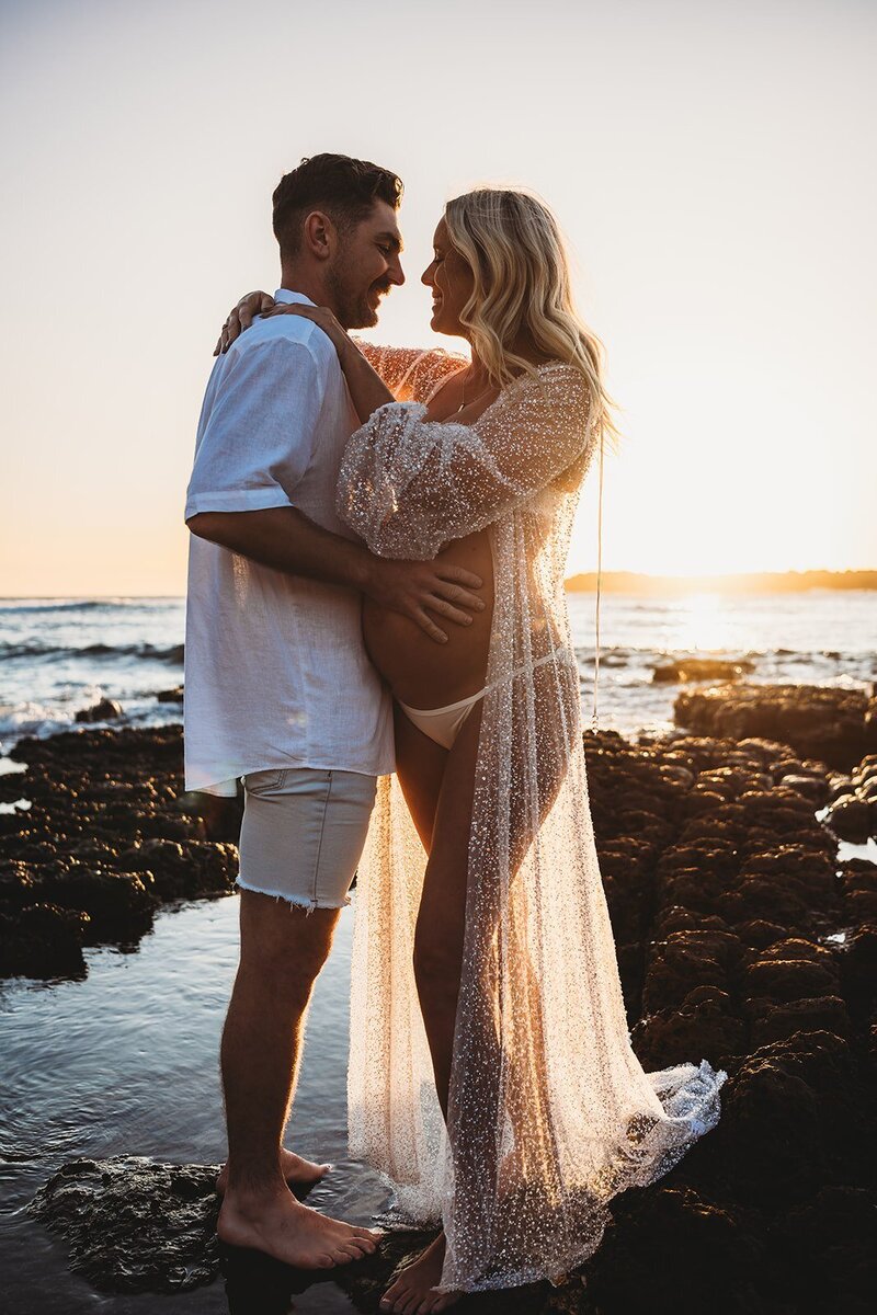 Pregnant woman and man embrace on the rocky shore at sunset during a stunning South Coast maternity session. She wears a sheer cover-up, and he dons a white shirt and shorts, captured beautifully by an NSW maternity photographer.