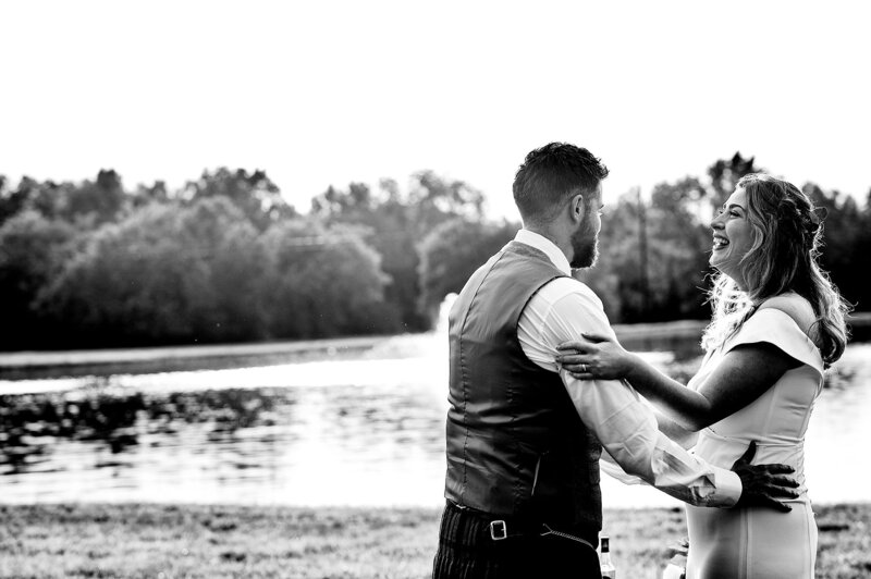 The groom dressed in a kilt and a vest embraces the bride who is laughing. The bride is wearing a fitted off the shoulder dress as they stand lakeside at Sugarfoot.