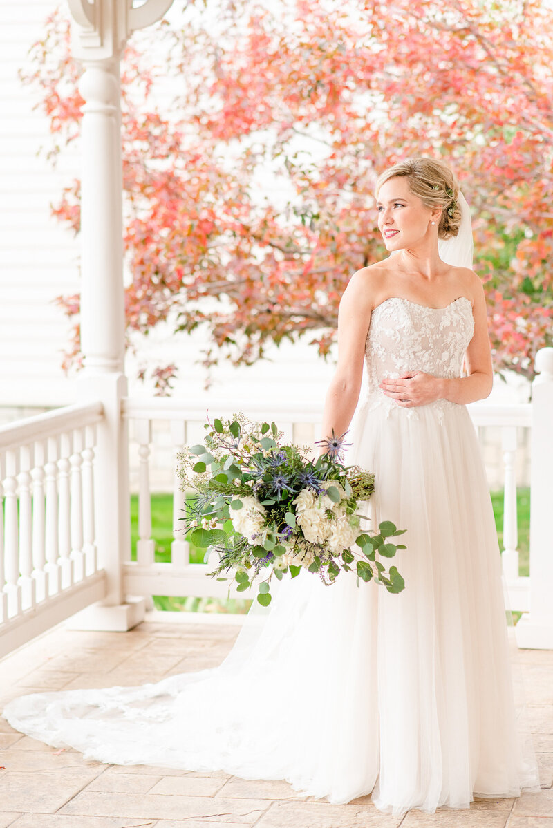 Full body portrait of a bride on the porch of a wedding venue with red tree in background.