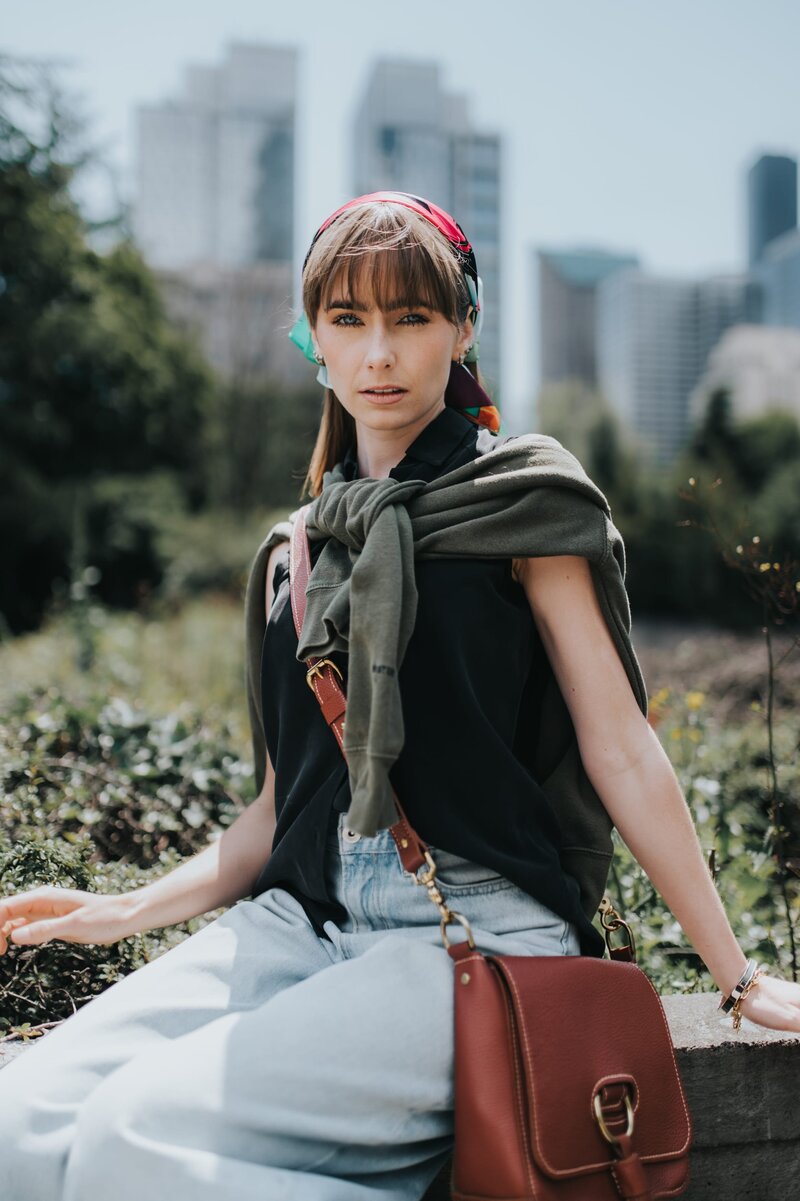 Woman with colored scarf in her and bangs wears a brown purse and a green sweater tied over her shoulders as she sits outside.