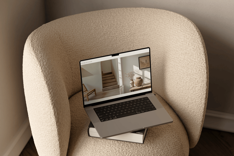 A beige boucle occasional chair holds a thick hardcover book with an open laptop resting on top. The screen displays the final design scheme for a virtual interior design project, marking the culmination of a thoughtfully curated client experience.