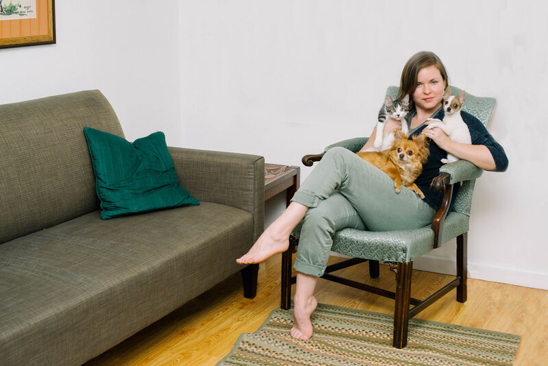 Michelle-Marie Gilkeson sits legs-crossed in a green chair holding two small dogs  and one kitten
