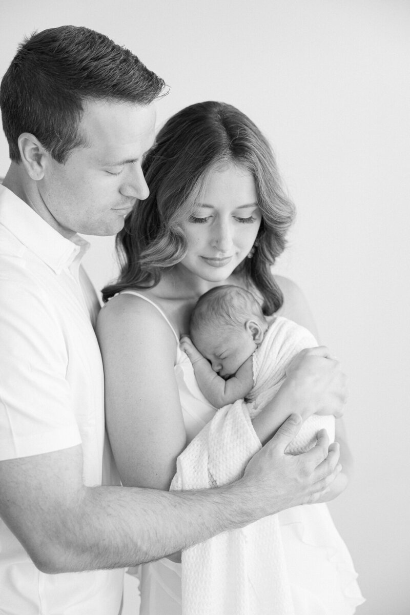 black and white image of a mother holding newborn baby curled against her chest while dad embraces her as they look that their child, Indianapolis newborn photographer