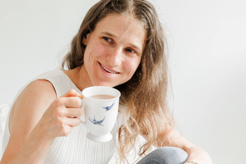 Woman sitting down, holding a cup of tea and smiling off to the side.