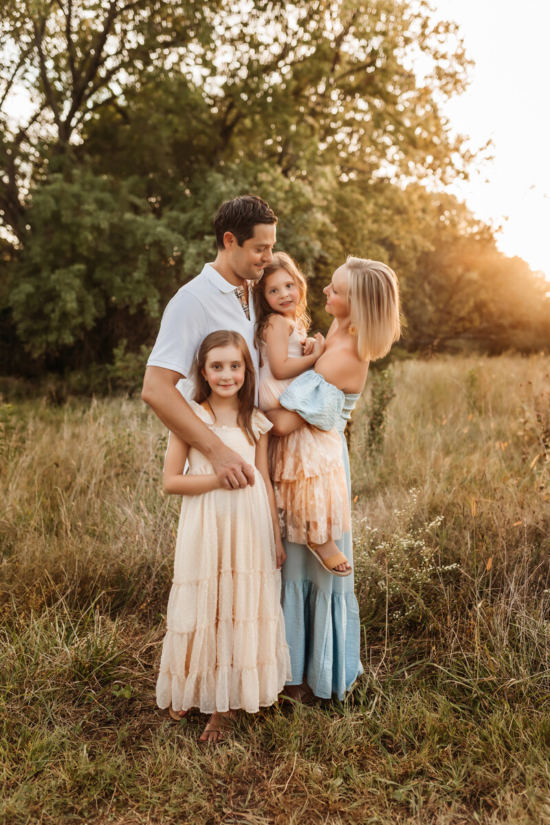 Two girls smile at Kansas City Mo photographer with their parents holding them in field at sunset family session.