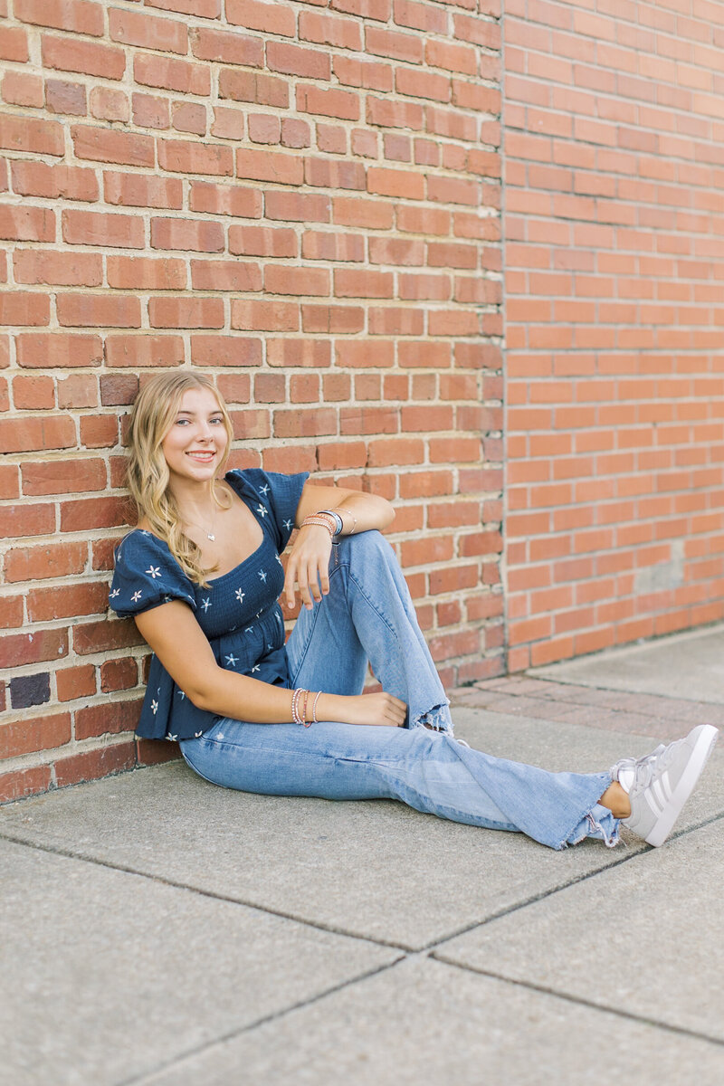 Senior photo of a high school girl sitting casually against a brick wall in downtown Raleigh, photographed by Raleigh Senior Photographer Lindsey Lambert Photography.