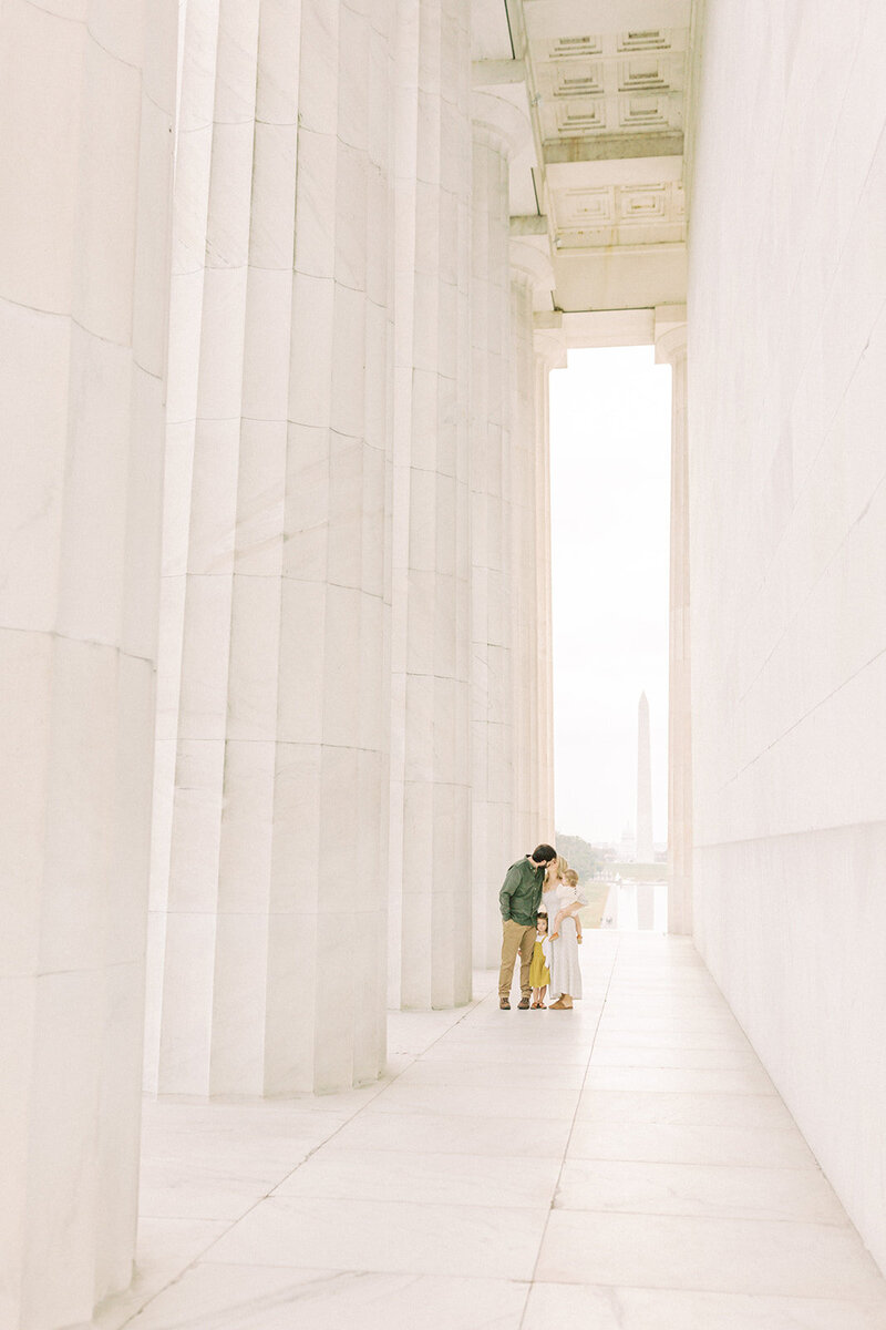 A mother and father stop and kiss one another while standing in the Lincoln Memorial with their two daughters during their photo session photographed by Washington DC Family Photographer Marie Elizabeth Photography