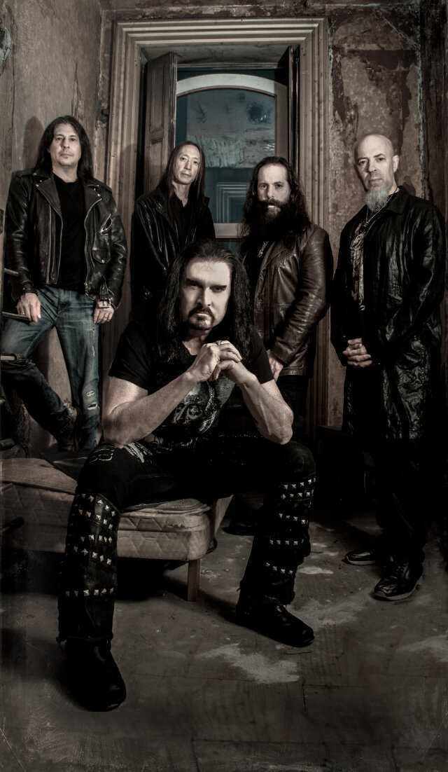 Music group portrait Dream Theater standing in old hotel room around singer sitting on bed