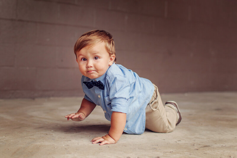 Crawling baby with a bowtie and blue shirt looking at the camera with a cute expression.