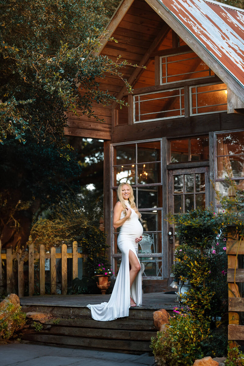 Blonde mom to be posing in front of a greenhouse at Out Under the Trees in Angleton, Texas.