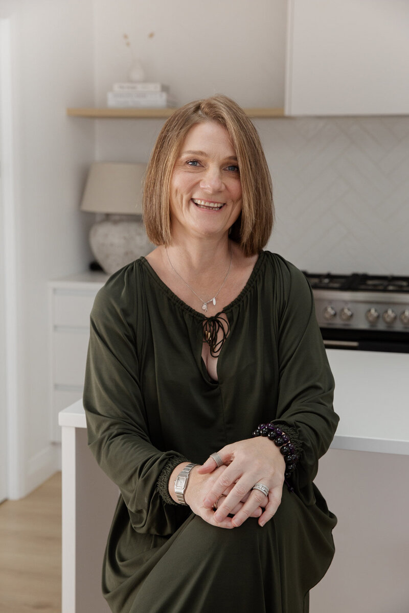 Portrait shot of energy healer Sharon Emery, sitting on a barstool in her kitchen.