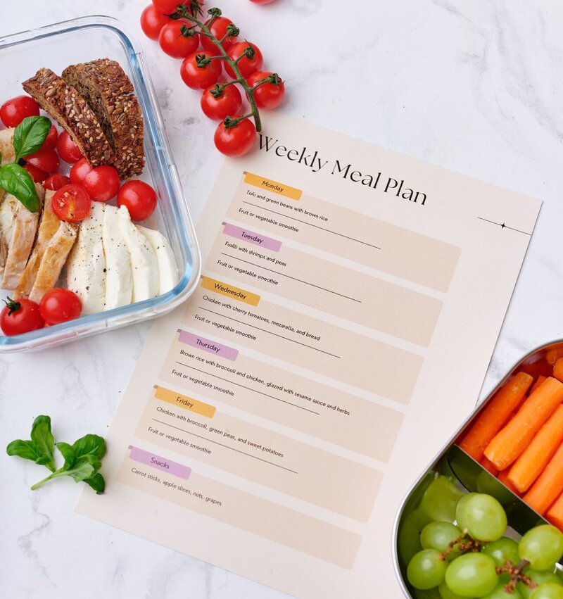 Midlife meal planning scene featuring a weekly meal plan sheet, sliced chicken, mozzarella, cherry tomatoes, seeded bread, and a snack box with carrots and grapes—part of the Menopause.Rescripted Toolkit for balanced nutrition and meal prep.