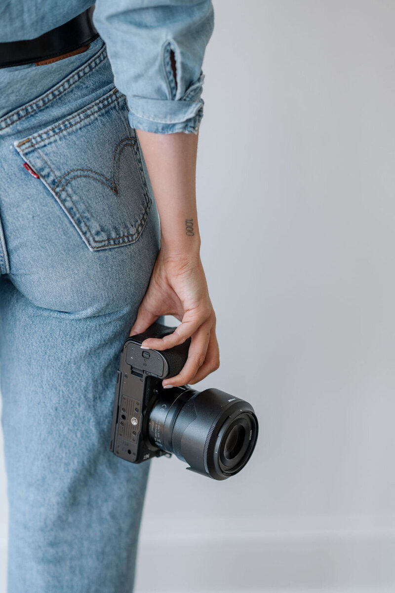 A close up of Emily holding her camera at her side. She is wearing light wash denim jeans and a matching denim button up.