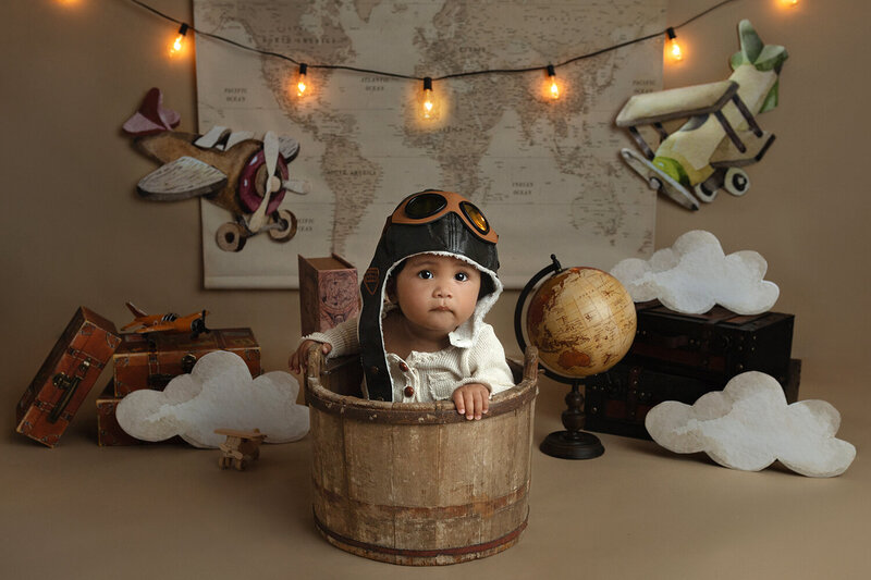 A toddler baby wears antique airman goggles while sitting in a bucket and decorated by a Lafayette Baby Milestone Photographer