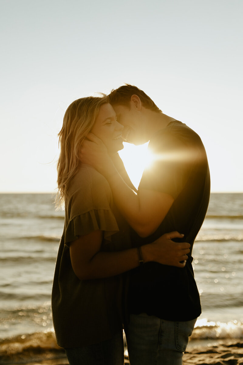 A man kissing his girlfriend on the cheek at the beach while the sun is setting