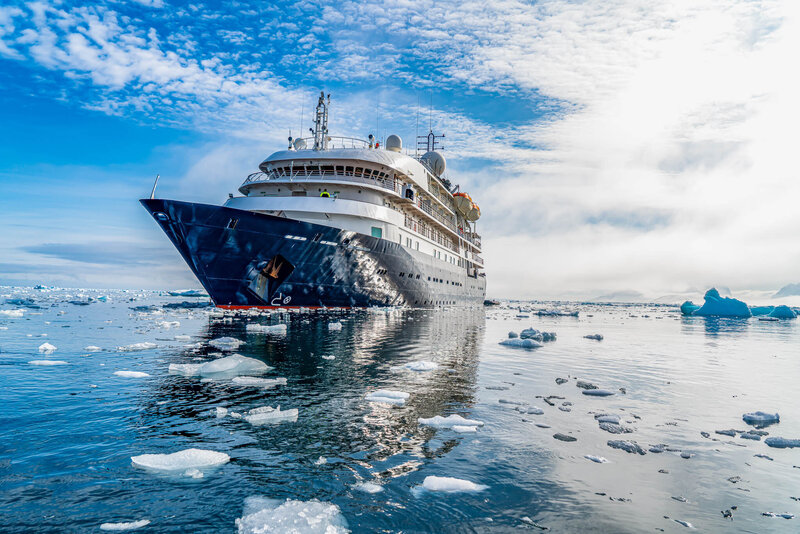 A large expedition cruise ship navigating icy waters surrounded by floating ice chunks under a partly cloudy blue sky.