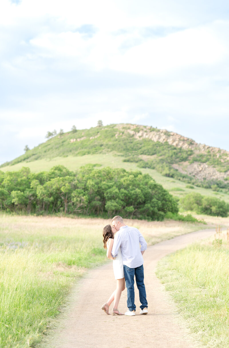 Couple going in for a kiss while standing on a dirt pathway with the mountains behind them