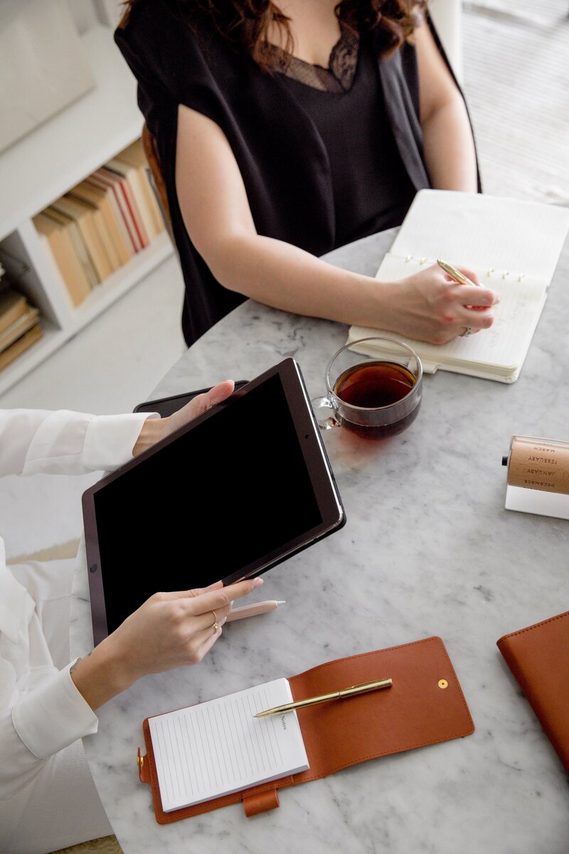 Woman writing in a notebook sitting next to a woman holding a tablet