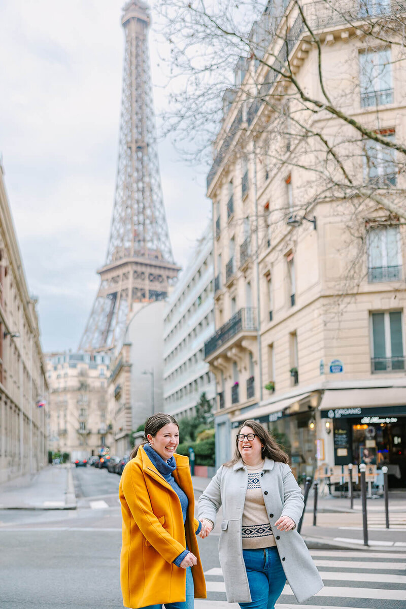 Two women laughing and holding hands while wearing jumpers and long colorful jackets during a Paris photoshoot near the Eiffel Tower on rue de l’Université.