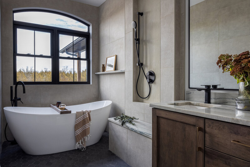 On the right is a freestanding tub beneach a black arched window. The hand shower is in a shower niche with a tiled bench. On the right is the oak vanity with marble counter top.
