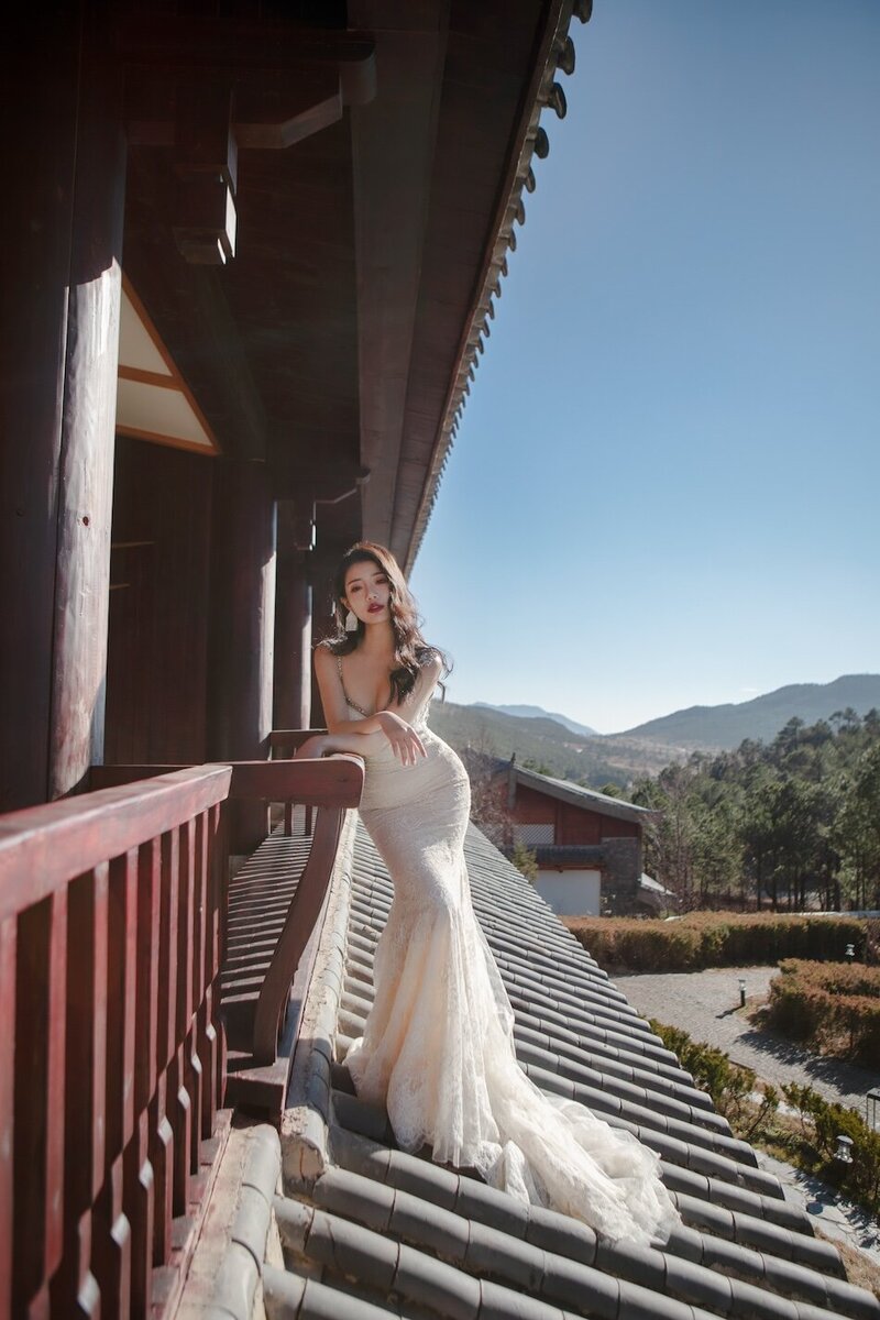 Bride in couture dress on a building awning leaning against a balcony rail.