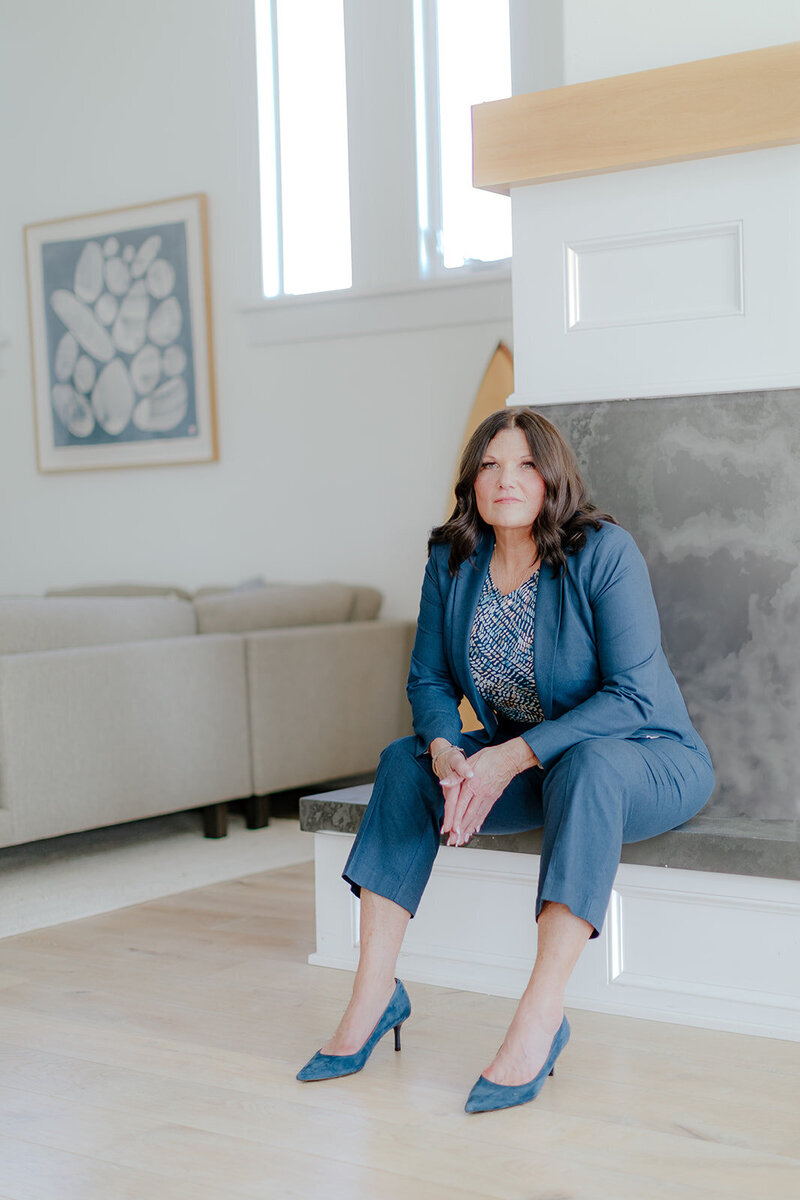 Professional full-body portrait of Susie Schumacher sitting on a modern fireplace ledge, wearing a navy pantsuit and heels, looking confident and composed in a bright, minimalist living space – Susie Schumacher Life Coach