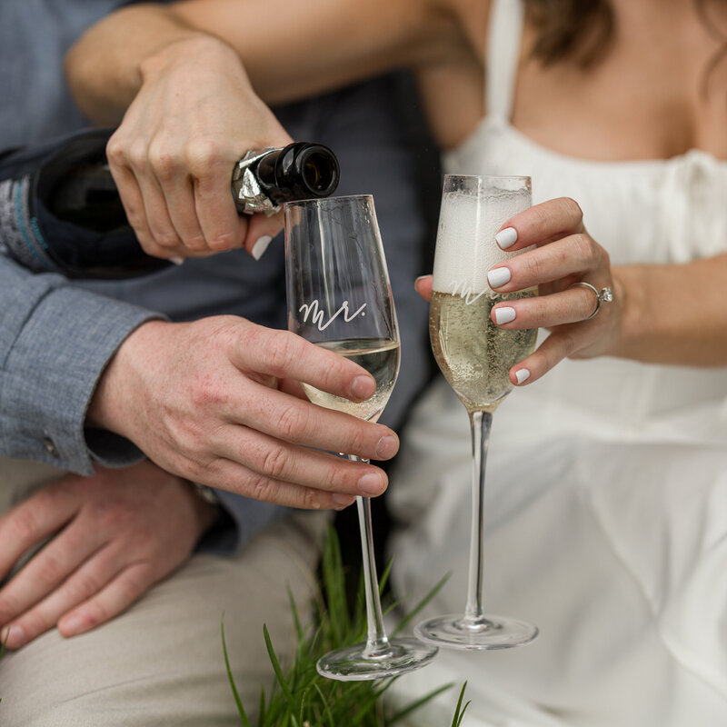 A closeup of hands pouring champagne into glasses during an engagement session, showcasing her sparkling engagement ring and celebratory elegance.
