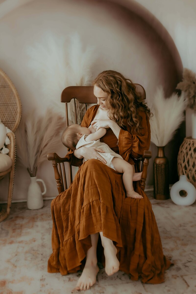 Woman in a brown dress sits in a rocking chair holding a baby. The room has bohemian decor with wicker furniture and soft lighting.