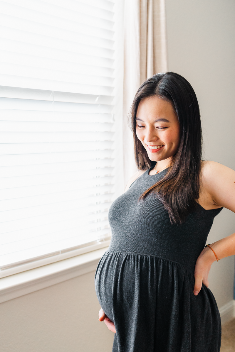 A pregnant woman dressed in a long charcoal dress stands by a window. She holds her belly with one hand while supporting her back with her other hand. She looks down at her belly and smiles softly in this sweet photo captured by an Austin Maternity Photographer
