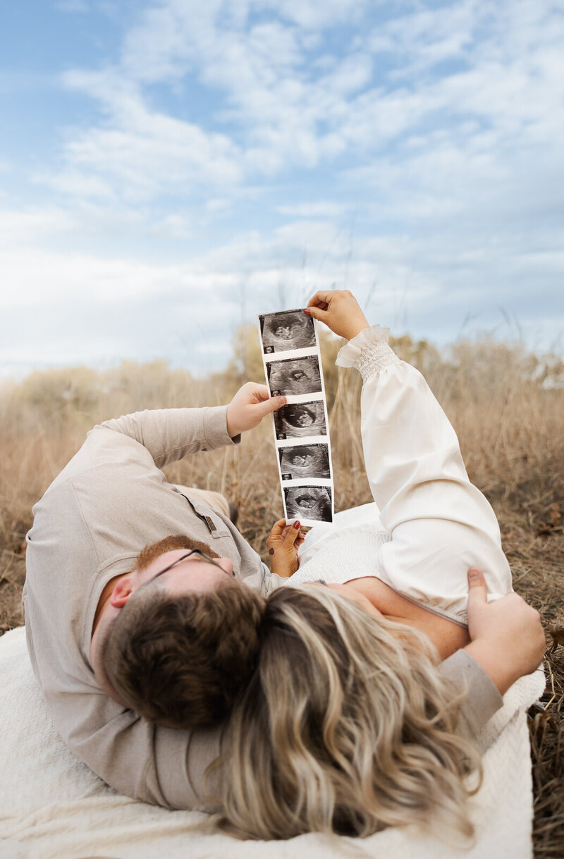 Expecting couple laying in a field on a blanket holding their ultrasound images up with a blue cloud filled sky in the background. 