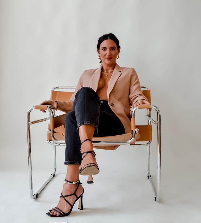 Emily Michele sitting confidently on a modern chair wearing a beige blazer and heels, embodying leadership, empowerment, and self-assured energy.