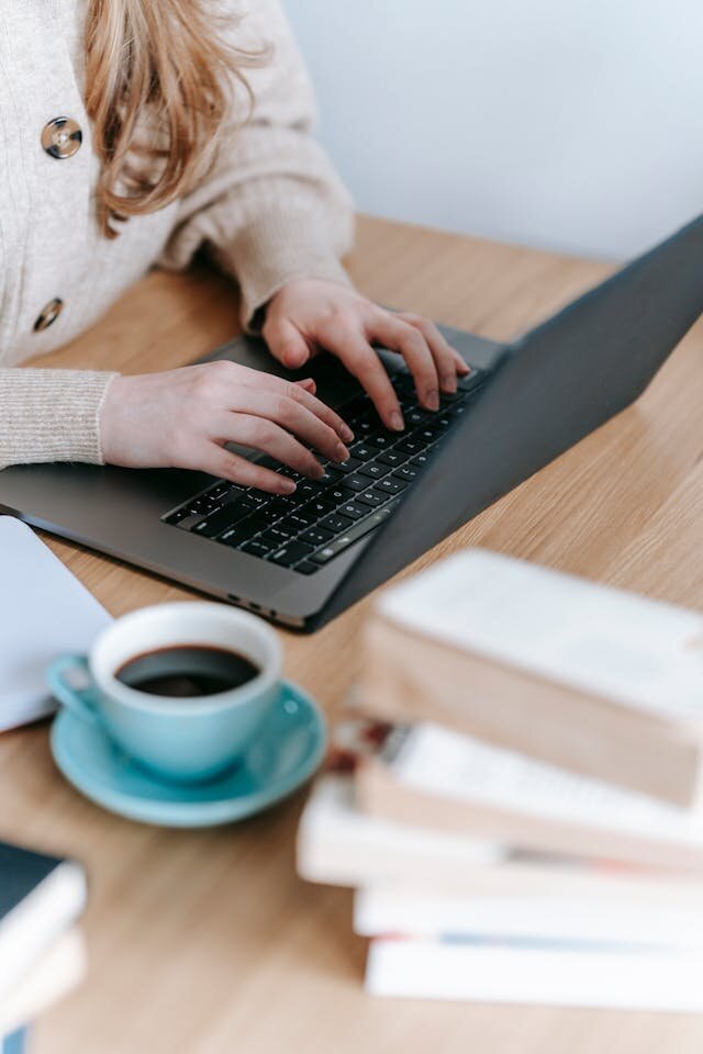 Gros plan sur une jeune femme qui tape à l'ordinateur sur son bureau, avec un café et des livres à côté d'elle