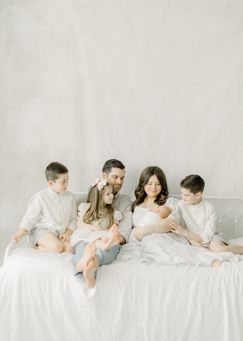 Beautiful Dallas family sitting on a bed together in a DFW photography studio while they are leaning in and as the siblings are looking at their newborn baby brother.
