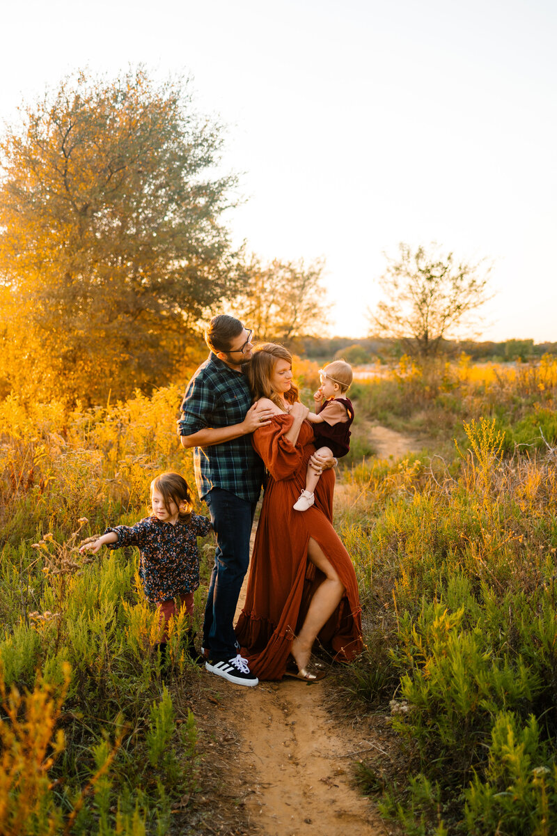 Family photograph in the forest, the woman is pregnant and has a long orange dress and is carrying a baby girl in a red dress. The man in a green plaid shirt is looking at the baby and behind him is a little girl standing and touching some flowers