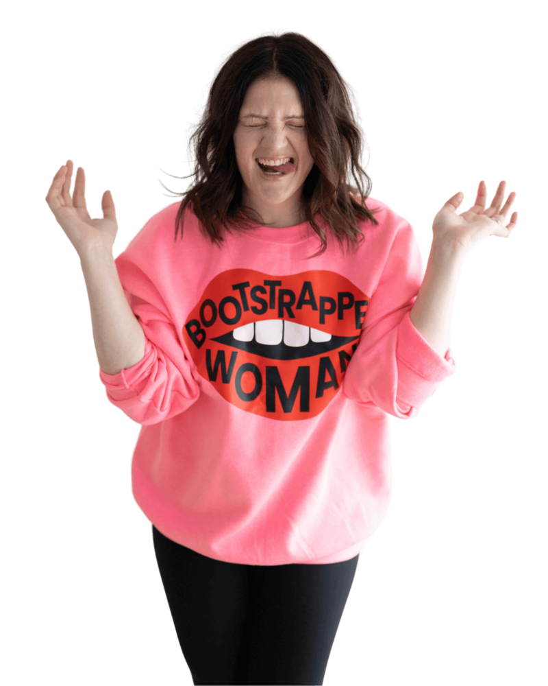 Emily closes her eyes and throws her hands in the air while sticking out her tongue and wearing a pink sweatshirt that says "bootstrapped woman."