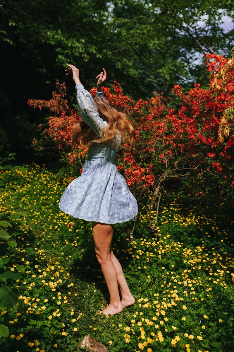 -Woman twirls in pale blue dress in a field of yellow flowers in front of tree with vibrant burgundy flowers.
