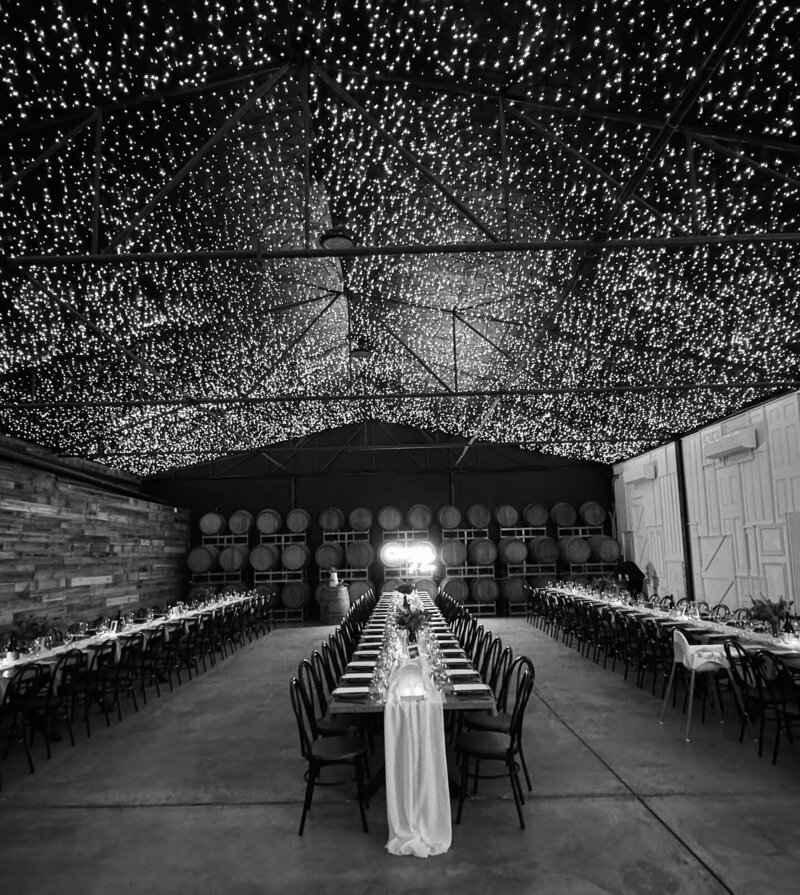 A black and white image of a rustic barn set up for a wedding reception with a wall of wine barrels and a ceiling full of twinking fairy lights