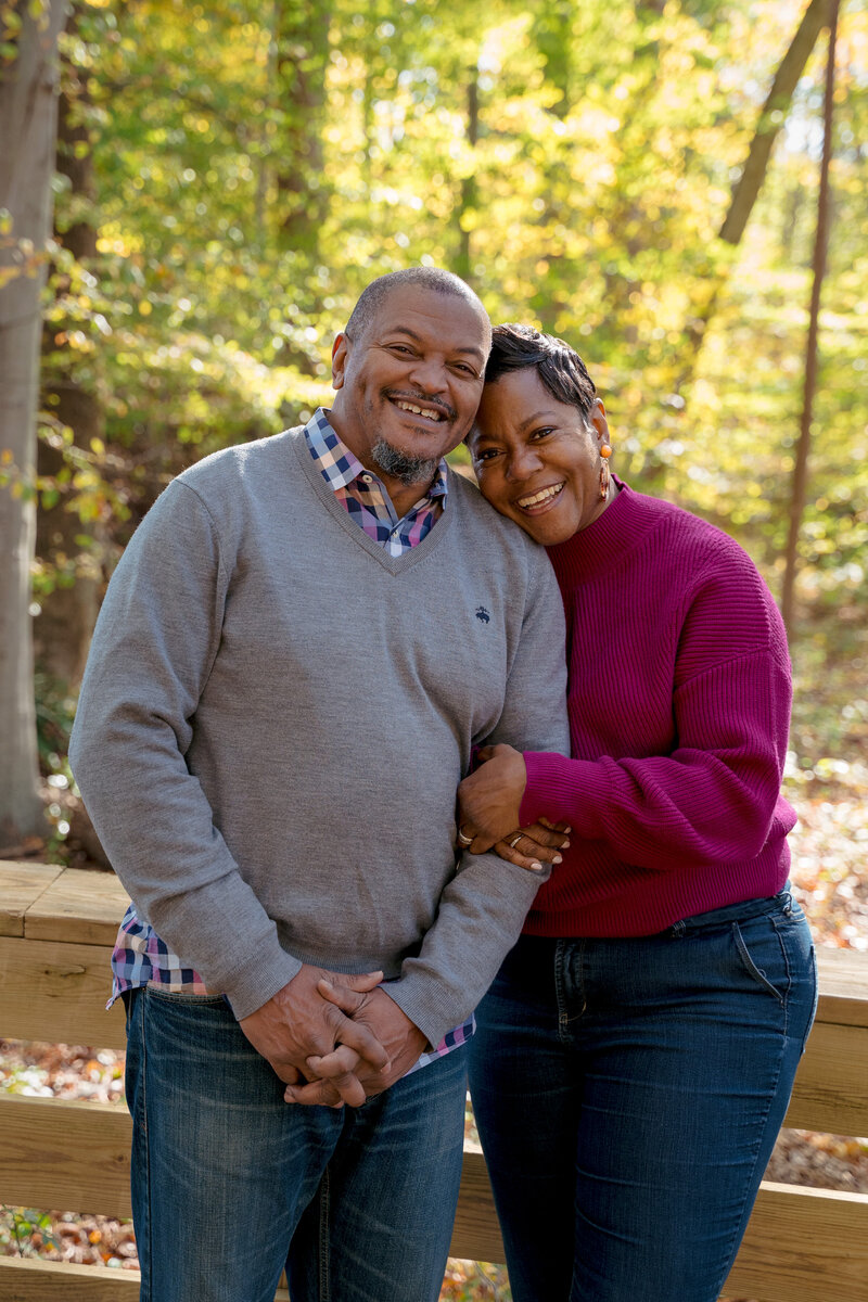 An older couple smiling in a park. One has their arms wrapped through the other's as they rest their head on their shoulder.