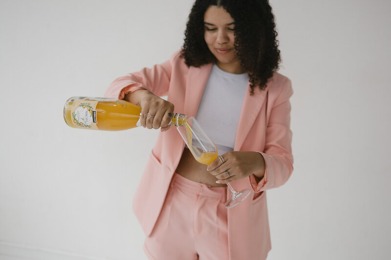 Photo of Catryce pouring mimosa into a glass in a pink jumpsuit on a neutral background