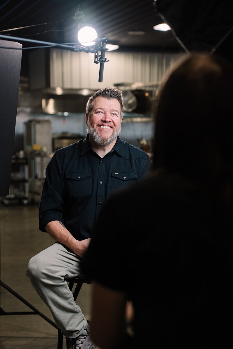 A man with short, salt-and-pepper hair and a beard smiles during an on-camera interview, seated on a stool in a well-lit industrial kitchen. A boom microphone hangs above him, and a crew member is visible in the foreground out of focus.