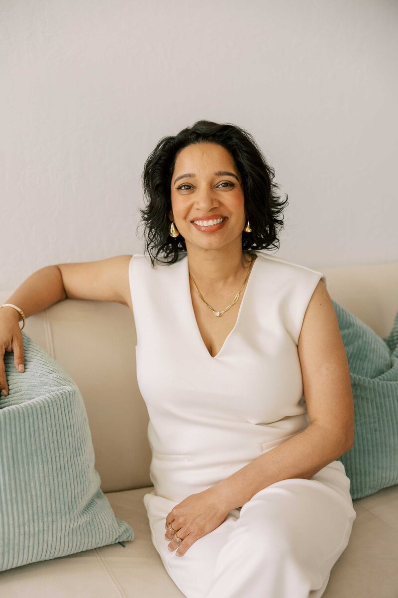 Woman leaning casually on a table, wearing an all white outfit and smiling at the camera