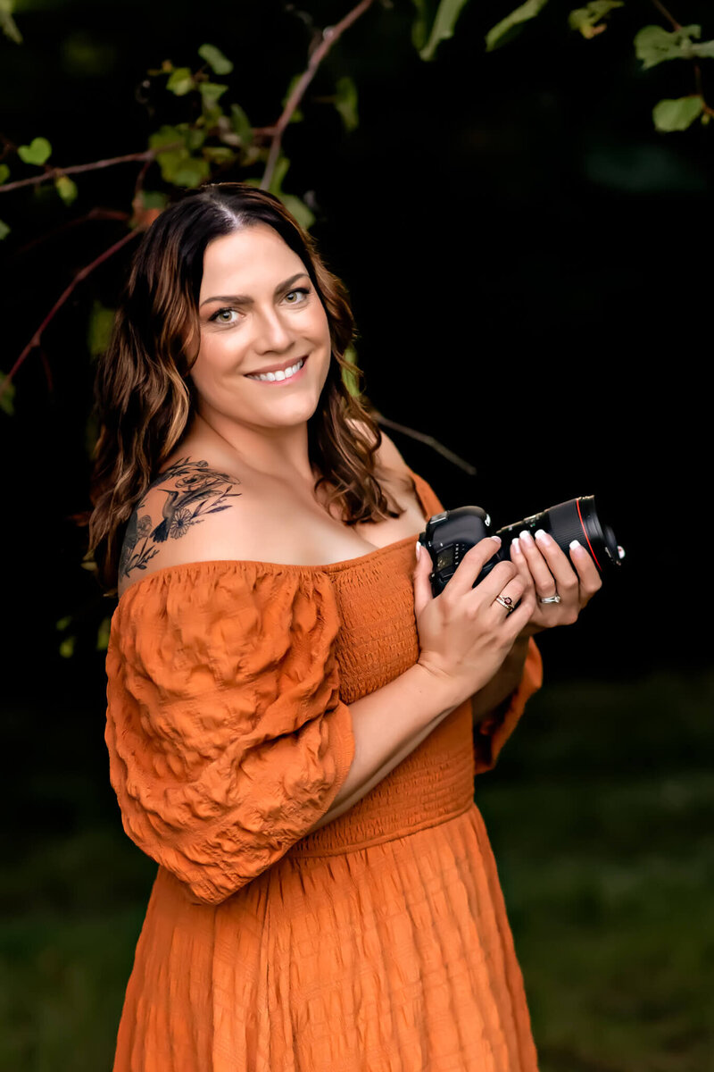 Woman wearing a long orange dress is standing in a field of tall wild grass. She is hugging onto herself and smiling.