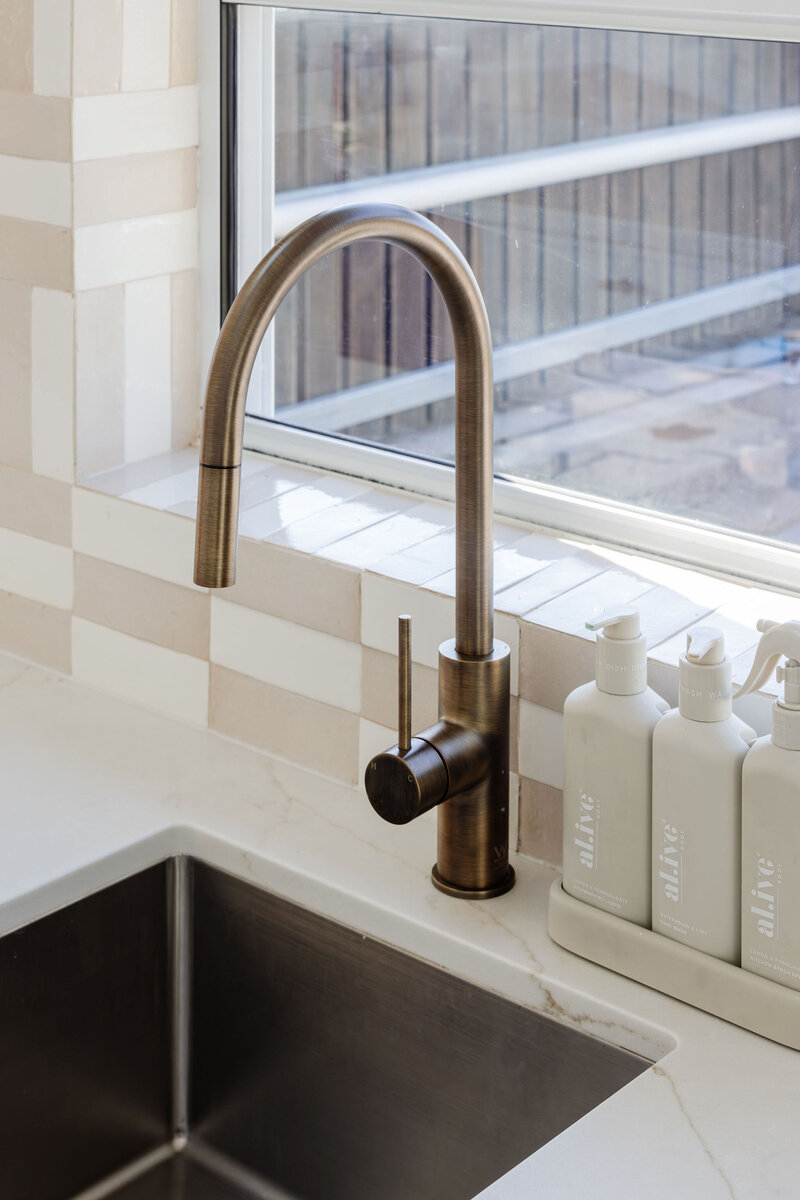 A brushed bronze kitchen faucet by a window complements the sink and soap dispensers on a white counter, embodying the elegance of luxury home design in Newcastle.