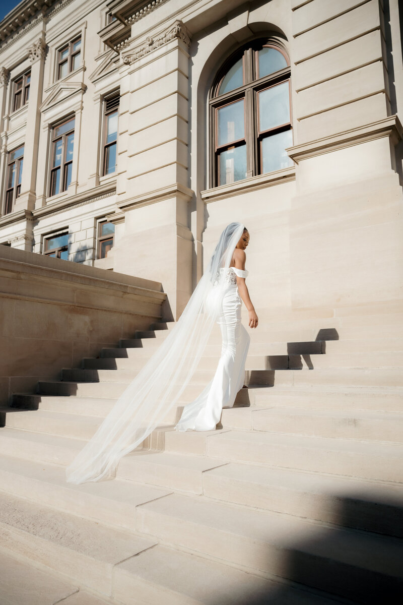 Elegant bride in an off-the-shoulder gown with a long veil ascending courthouse steps, framed by classic architecture and soft natural light.