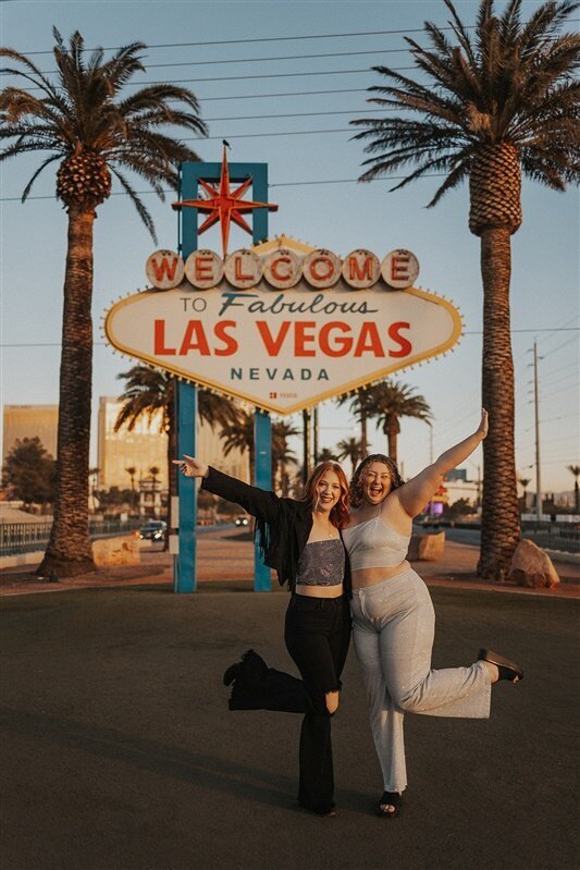 Steph and Andi pose cheerfully in front of the iconic "Welcome to Fabulous Las Vegas, Nevada" sign, each raising an arm and lifting a leg in a playful manner. Palm trees and the early evening sky frame the background.