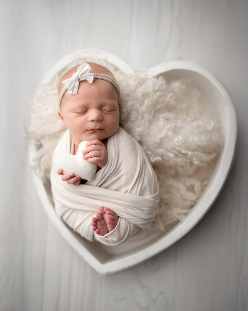 Newborn Baby girl sleeping posed in a white heart bowl prop and holding a small white heart